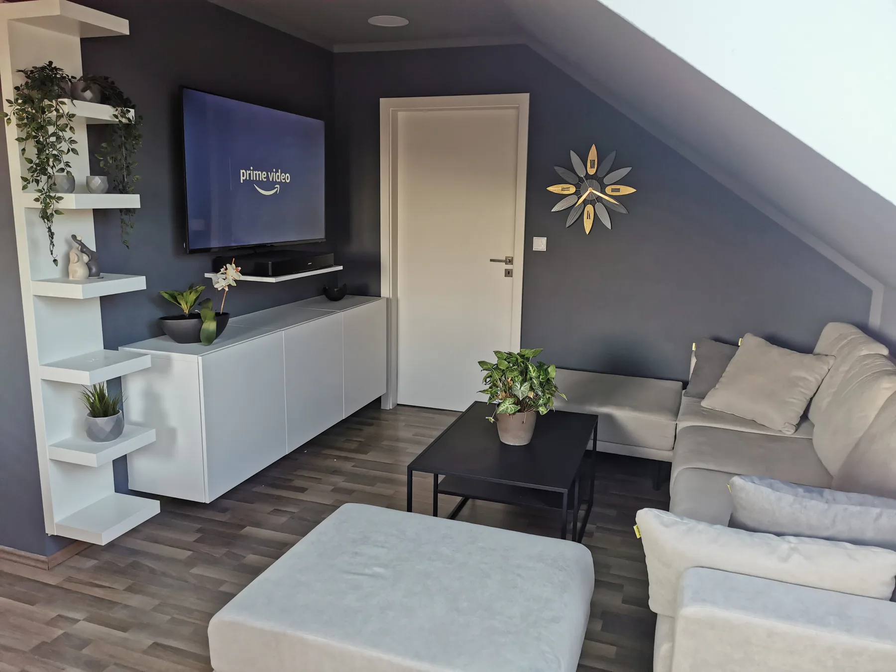 Wohnzimmer mit heller Eckcouch, Hocker und großem Smart-TV